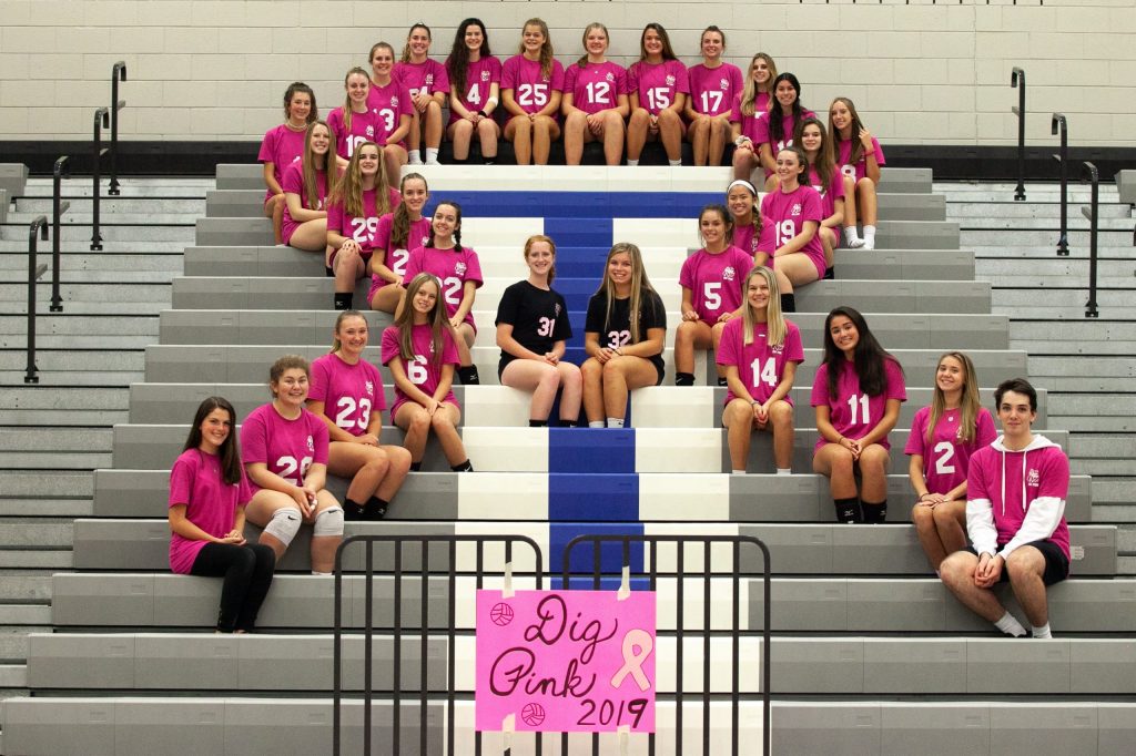 Team members sit on the school bleachers in a breast cancer ribbon formation.